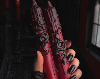 May include: Two deep red taper candles featuring a raised black snake relief design and decorative wax drips running down the sides. The candles have a smooth, matte finish and are cylindrical in shape, showcasing a detailed serpent motif wrapped around the center of each candle body.