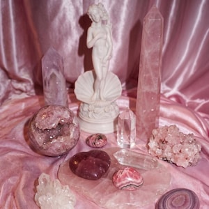 Peut inclure: Une collection de divers cristaux et pierres comprenant une géode sphérique, une grande pointe de quartz rose, une pointe de quartz clair, une pierre en forme de cœur, une agate rubanée et une figurine blanche représentant une personne dans un coquillage.