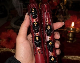 May include: Two deep red taper candles featuring a vertical row of three-dimensional black skull embellishments. Each skull is accented with small gold-toned floral details and tiny red rhinestones, with wax drips running down the candle surface.