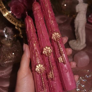 May include: Three tall, cylindrical taper candles in a deep rose-pink color, featuring a textured, dripped wax finish. Each candle is adorned with a metallic gold-toned decorative key emblem attached to the front surface of the wax.