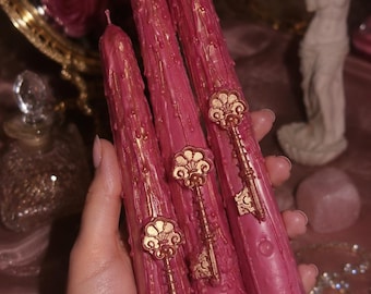 May include: Three tall, cylindrical taper candles in a deep rose-pink color, featuring a textured, dripped wax finish. Each candle is adorned with a metallic gold-toned decorative key emblem attached to the front surface of the wax.