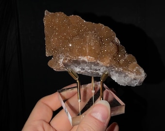 May include: A brown, crystalline mineral specimen with a textured, sparkling surface. The mineral is mounted on a clear, square acrylic base supported by three thin, gold-toned metal prongs that hold the specimen securely in place.