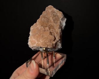 May include: A rough, light brown mineral specimen with a crystalline, sparkling surface texture. The mineral is mounted on a clear acrylic square base supported by three thin, vertical gold-toned metal prongs that hold the stone upright.