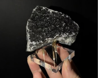 May include: A raw, dark black mineral specimen with a crystalline, textured surface. The stone is mounted on a clear square acrylic base supported by three thin, vertical gold-toned metal rods that hold the mineral securely in place.