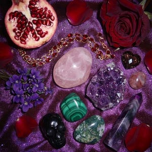 May include: A collection of crystals, gemstones, and floral elements on a purple, glittery surface. Includes a rose quartz stone, amethyst cluster, malachite, a black skull, a pomegranate, and a red rose.
