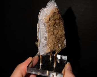 May include: A vertical slice of brown and white agate geode with a crystalline texture, mounted on a clear square acrylic base with three gold-toned metal prongs that support the mineral specimen upright against a dark background.