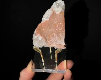 May include: A triangular-shaped mineral specimen featuring a textured, pale peach-colored crystalline surface bordered by translucent white quartz edges, mounted on a clear acrylic stand with two gold-toned metal support prongs.