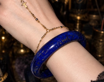 May include: A gold-toned slave bracelet featuring a delicate chain connected to an eye-shaped ring and a central ankh symbol, paired with a thick, polished deep blue lapis lazuli bangle bracelet with visible gold-colored mineral flecks.