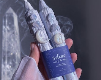 May include: Two light blue taper candles with a textured, dripped wax finish. Each candle features embedded iridescent moon and star charms and small polished stones. A dark blue paper band wraps around the center with the text Selene Lady of the Moon.