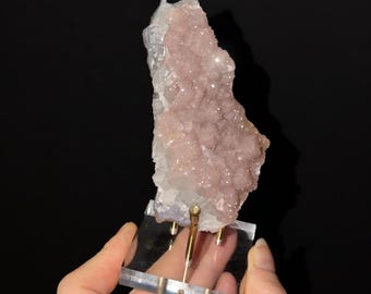 May include: A vertical mineral specimen featuring a cluster of pale pink, crystalline formations attached to a translucent white base. The mineral is mounted on a clear acrylic stand with a gold-toned metal support rod for display.