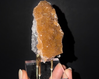 May include: A raw, amber-colored druzy crystal cluster with a sparkling, textured surface. The mineral specimen is mounted vertically on a clear acrylic base supported by three thin, gold-toned metal rods for display purposes.