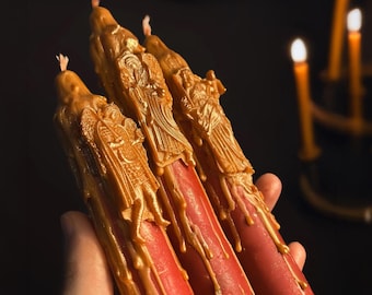 May include: Three red taper candles featuring gold-colored wax relief carvings of winged figures. The gold wax appears to drip down the sides of the smooth red cylindrical bodies. Each candle is topped with a small, unlit wick at the pointed end.