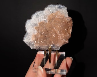 May include: A raw mineral specimen featuring a central cluster of light brown, sparkling crystals surrounded by a jagged, translucent white crystalline border, mounted on a clear acrylic base with three thin gold-toned metal support rods.