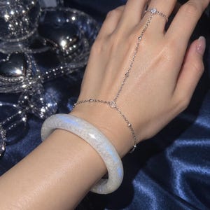 May include: A silver-toned hand chain featuring a delicate link design with small beads and a single ring component, paired with a thick, translucent white bangle bracelet that displays a subtle iridescent blue sheen across its smooth surface.