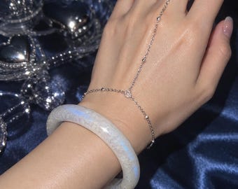 May include: A silver-toned hand chain featuring a delicate link design with small beads and a single ring component, paired with a thick, translucent white bangle bracelet that displays a subtle iridescent blue sheen across its smooth surface.