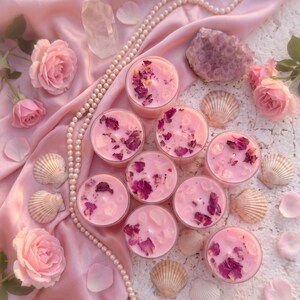 May include: Pink candles with dried rose petals and crystals are arranged on a pink satin cloth, surrounded by roses, seashells, and a pearl necklace. A clear crystal and a purple geode are also present.