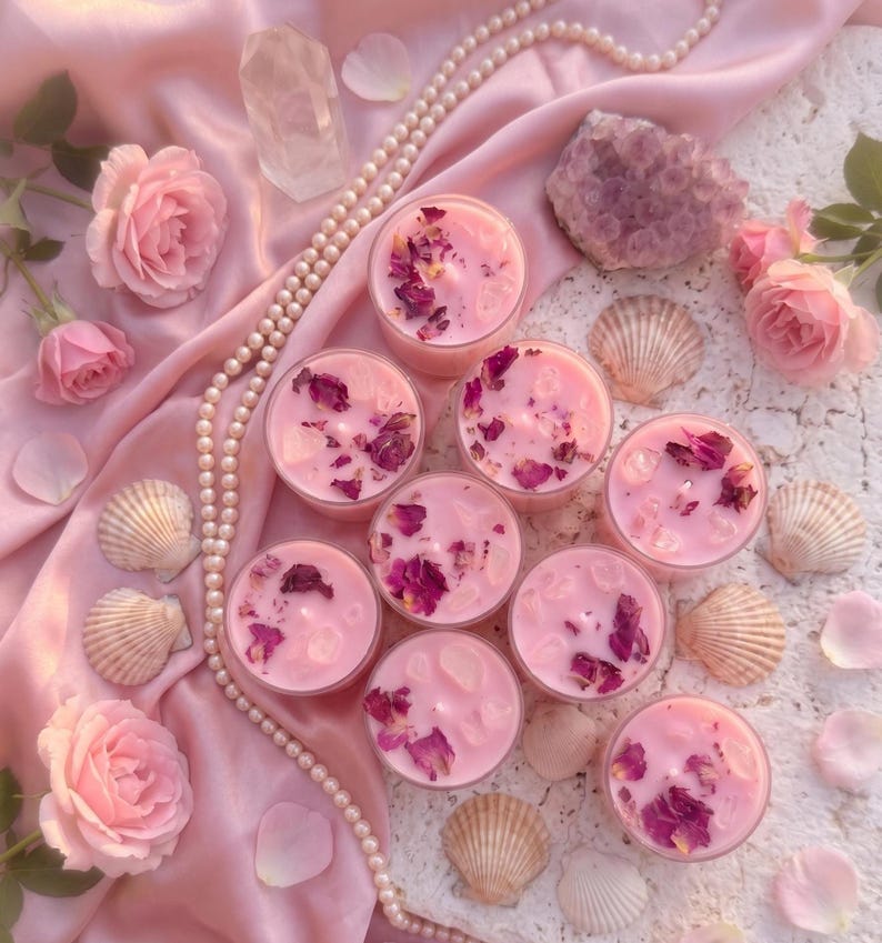 May include: Pink candles with dried rose petals and crystals are arranged on a pink satin cloth, surrounded by roses, seashells, and a pearl necklace. A clear crystal and a purple geode are also present.