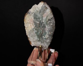 May include: A raw mineral specimen featuring a central cluster of small, sparkling green crystals surrounded by jagged, translucent white and pale beige rock formations, mounted on a clear acrylic base with gold-toned metal prongs.