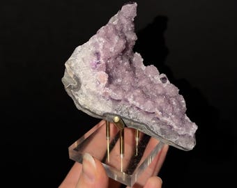 May include: A jagged amethyst crystal cluster with light purple, crystalline textures. The specimen is mounted on a clear, square acrylic base supported by three thin, vertical gold-toned metal rods that hold the stone in an upright position.