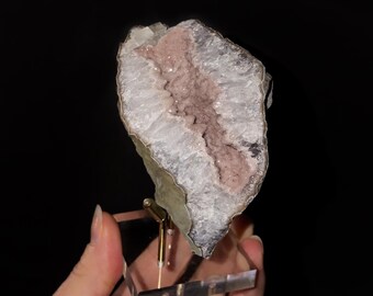 May include: A raw mineral specimen featuring a central band of dusty pink crystals surrounded by translucent white quartz formations, mounted on a clear acrylic base with a gold-toned metal support stand against a black background.