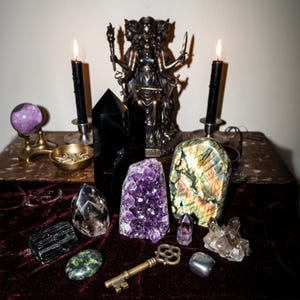 May include: A collection of crystals and ritual items including a bronze three-faced goddess statue, two black candles in silver holders, a purple sphere, a black obsidian tower, an amethyst cluster, a labradorite slab, and a brass key.