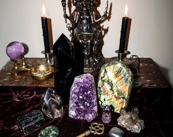 May include: A collection of crystals and ritual items including a bronze three-faced goddess statue, two black candles in silver holders, a purple sphere, a black obsidian tower, an amethyst cluster, a labradorite slab, and a brass key.