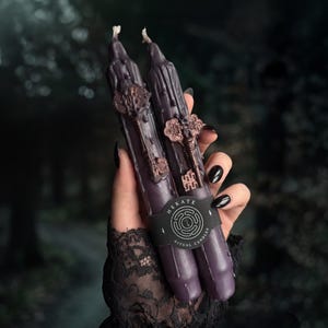May include: Two dark purple taper candles with a dripped wax texture, each featuring an attached metallic copper-colored skeleton key charm. A black paper band wraps around the center, printed with the text Hekate Ritual Candles.