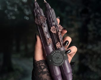 May include: Two dark purple taper candles with a dripped wax texture, each featuring an attached metallic copper-colored skeleton key charm. A black paper band wraps around the center, printed with the text Hekate Ritual Candles.