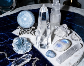 May include: A collection of crystals including a light blue sphere on a silver stand, a clear quartz tower, a white selenite tower, a blue celestite geode, a moonstone, a small blue tumbled stone, a selenite wand, and a clear crystal crescent moon.
