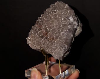 May include: A cluster of light purple, crystalline mineral formations with a textured, sparkling surface. The specimen is mounted on a clear, square acrylic base supported by two thin, vertical gold-toned metal prongs.