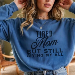 Puede incluir: Una sudadera azul con el texto "TIRED Mom BUT STILL GIVING MY ALL" en letras negras. La sudadera es de color azul liso y tiene mangas largas.