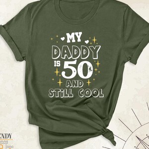 May include: Olive green t-shirt with the text "MY DADDY IS 50 AND STILL COOL" in white. The design includes hearts and stars. The shirt is knotted at the bottom.