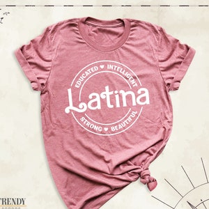 May include: A pink t-shirt with a white circle graphic that says "Latina" with the words "Educated", "Intelligent", "Strong", and "Beautiful" around the circle.