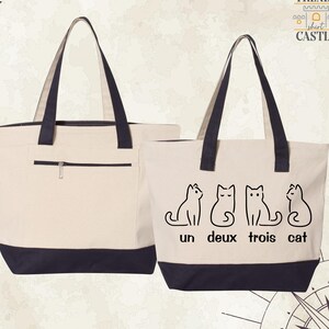 May include: Two beige tote bags with black handles and black bottoms. One bag has a zipped pocket on the front. The other bag has a graphic of four cats with the text "un deux trois cat" printed on the front.