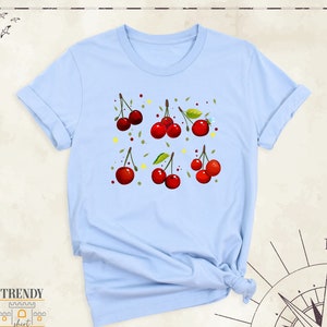 May include: Light blue t-shirt featuring a pattern of red cherries and green leaves. The cherries are arranged in a grid pattern on the shirt.