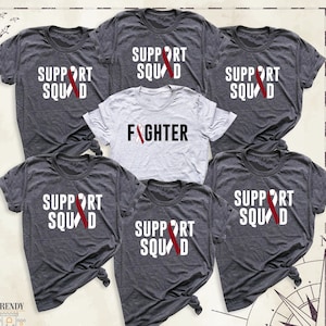 May include: Six grey t-shirts with white text and a red ribbon design. One shirt says "Fighter" with a red ribbon. The other five shirts say "Support Squad" with a red ribbon.