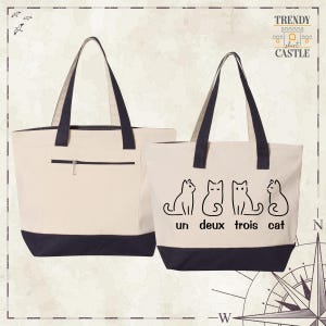 May include: Two beige tote bags with black handles and black bottoms. One bag has a zipped pocket on the front. The other bag has a graphic of four cats with the text "un deux trois cat" printed on the front.