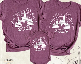 May include: Two maroon short-sleeve t-shirts and one maroon short-sleeve baby bodysuit. Each garment features a white graphic of a castle silhouette, a mouse ear outline, fireworks, stars, and the year 2026 printed in a bold font.