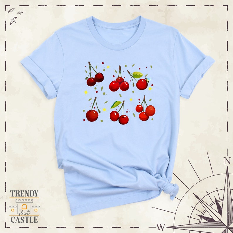 May include: Light blue t-shirt featuring a pattern of red cherries and green leaves. The cherries are arranged in a grid pattern on the shirt.