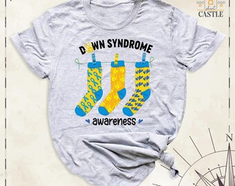 May include: A light gray short-sleeve t-shirt featuring a graphic of three socks hanging on a clothesline. The text reads DOWN SYNDROME AWARENESS. The socks are yellow and blue with patterns of ribbons, handprints, and hearts.