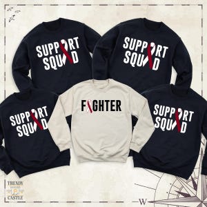 May include: Set of four navy blue and cream sweatshirts with red ribbon designs. The sweatshirts feature the text "Support Squad" and "Fighter".