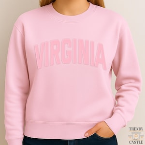 May include: A light pink crewneck sweatshirt featuring the word VIRGINIA in a large, arched, tonal block-letter font across the chest. The garment has long sleeves with ribbed cuffs and a matching ribbed hemline at the waist.