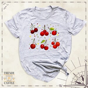 May include: A grey t-shirt featuring a design of red cherries and green leaves. The cherries are scattered across the shirt in a random pattern.