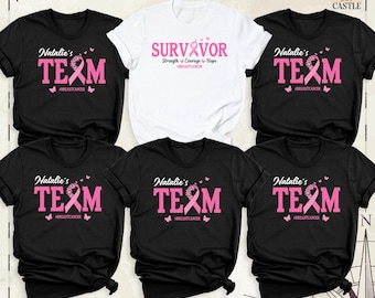 May include: Five short-sleeved t-shirts arranged in a group. Four are black with pink text reading Natalie's TEAM and #BREASTCANCER with a pink ribbon. One white t-shirt in the center features pink text reading SURVIVOR Strength Courage Hope.