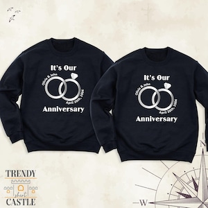 May include: Two navy blue crewneck sweatshirts featuring a white graphic of two interlocking rings with a diamond. The text reads It's Our Anniversary, Olivia & John, and April 20th, 2004. The design is centered on the chest of each garment.