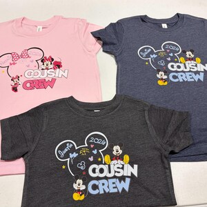 Puede incluir: Tres camisetas con gr&aacute;ficos de dibujos animados. Una camiseta rosa presenta a Minnie Mouse y las palabras "Cousin Crew". Una camiseta azul marino y una camiseta gris carb&oacute;n tienen a Mickey Mouse y las palabras "Cousin Crew".