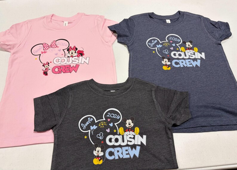 Puede incluir: Tres camisetas con gr&aacute;ficos de dibujos animados. Una camiseta rosa presenta a Minnie Mouse y las palabras "Cousin Crew". Una camiseta azul marino y una camiseta gris carb&oacute;n tienen a Mickey Mouse y las palabras "Cousin Crew".