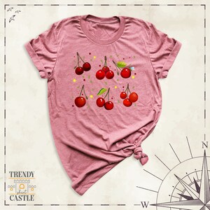 May include: A pink t-shirt featuring a design of red cherries on green stems with green leaves. The cherries are scattered across the shirt.