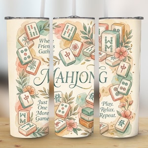 May include: A beige tumbler with a watercolor design featuring Mahjong tiles, flowers, and foliage. The text "MAHJONG" is prominently displayed, with additional phrases like "When Friends Gather" and "Play. Relax. Repeat."