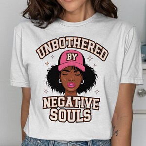 Può includere: Maglietta bianca con una grafica di una donna nera che indossa un berretto da baseball rosa con il testo "UNBOTHERED BY NEGATIVE SOULS".
